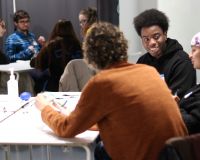 Three people sit around a table. There are more people in the background at another table. The table is white and the people are having a fun conversation