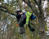 An image of Cheryl Duerden doing a woodland, aerial rescue drill, wearing a harness and is suspended in mid air, held up by ropes.