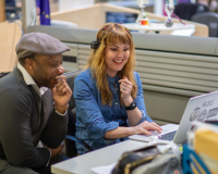 Two people sit on chairs at one of the hot desks at Pervasive Media Studio. They both look towards a laptop that sits on the desk smiling.