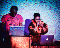 an image of two people looking at their laptops, one is speaking into a microphone, psychedelic visuals are project over them and the wall behind 
