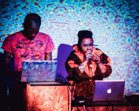 an image of two people looking at their laptops, one is speaking into a microphone, psychedelic visuals are project over them and the wall behind