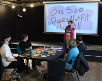 Still from a film of Graham speaking to 4 people sat at a table. A presentation says One Size Does Not Fit All in the background