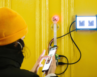 Photo of someone wearing a yellow hat, taken from behind, in front of a yellow door holding a mobile phone connected to another screen on the door