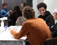 Three people are in the forefront of the image, sat around a table having a conversation. Another two people are in the background of the image, also sat around a table having a discussion.