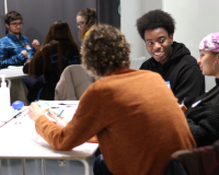 Three people are in the forefront of the image, sat around a table having a conversation. Another two people are in the background of the image, also sat around a table having a discussion.