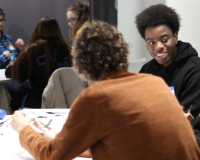 Two people are in the forefront of the image, sat around a table having a conversation. Another two people are in the background of the image, also sat around a table having a discussion.