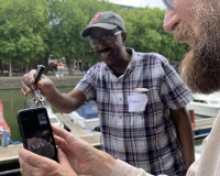 Two men standing by the harbour. one is holding up a mobile phone and another is shaking a set of keys in front of the phone.