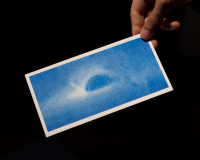 A hand holding a baby blue postcard depicting a black hole, against a black background.