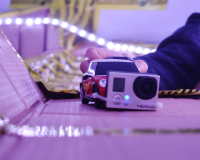 A child's hand places a remote control car with a go pro camera attached to the front of it onto a self built cardboard racetrack held together by tape with led strip lights attached in a DIY manner