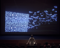 A man, shot from behind, experiences asses.masses in a theatre - the screen is awash with pixellated blue shapes. 