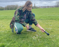 A man with shoulder length brown hair and a green jacket crouches in a grassy field holding a microphone towards the ground as if recording the sound of it