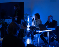 An image of a tranquil space, with an audience to the left, and three sound artists to the right, sat at a table, sharing a work of sound art. The room is lit in a peaceful, dark blue.
