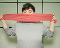 An image of a person, eyes closed, wearing a grey jumper, in front of a tiled background. They are stretching a piece of red rubber over their open mouth, creating an impression in the rubber.