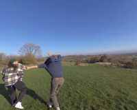 A screen capture from a 360 image of two people playfully moving around in a lush green space with a clear blue sky. 