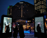 An image of a city at night, with two light fixtures in the foreground, on which human figures are silhoutted, interacting with the fixtures as part of a sensory exhibition. 