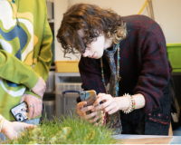 An image of a person examining an exhibit with their phone, intently. The exhibit is at the bottom of the picture, and is some kind of small grass installation. 