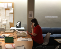 An image of Frances Bossom in her 90's style office installation at the arnolfini. She sits at a table, with an old corded phone to her left ear - on the table is a fax machine, and various pieces of paperwork. The wall in the background has a noticeboard covered in paper notices. 