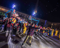An image of a large group of people of diverse physical needs, outdoors in an urban space, taking part in a collective theatrical experience; there is an installation behind the group. The people are smiling and contented.