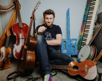 An image of the artist and creative, Greg Ryan, a white man in his mid to late 30's, sat amongst a vast array of predominantly acoustic instruments. He is looking directly into the camera, and wears blue jeans and a navy blue t shirt.