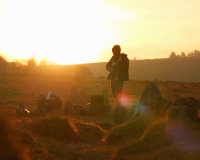 An image of artist Jim Wheale, playing guitar in the middle of a stone circle on the moor, at sunrise. He is silhouetted against a warm, yellow glow as he plays. 