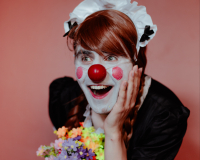 An image of Alistair Aitcheson in an androgynous clown/maid costume; he is cupping his head with his left hand with a bashful, cheery expression.