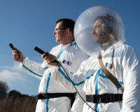 An image of two men, side by side, dressed in white uniforms that resemble astronaut uniforms. The man on the right, musician Mr. Kit, wears a fishbowl-like helmet on his head. Both men are extending their right arms outward, and are holding microphones with which to record their environment. 