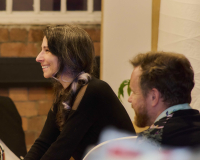 Two white people are smiling over to the left of the screen, they're both sat down.