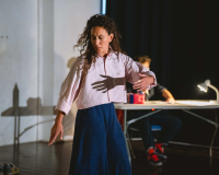 A woman dancing, mid turn, her long curly hair still spinning with the momentum. She wears a pink shirt and loose blue trousers. The performer from the last image out of focus behind her. Shadows on the wall.