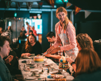 An image of Tom Marshman's tea party - Tom is standing, looking off camera to the right, about to cut a rainbow coloured cake sat on a white table, surrounded by happy participants.