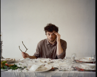 The artists sits at a messy seder table with glasses in one hand and hand in another