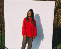 An image of a Emmanuella Blake-Morsi, against a white backdrop situated in an outdoor green space. Emmanuella is a black woman, wearing glasses, a red sweater and dark green trousers, looking off camera to the left, with a smile on her face. 