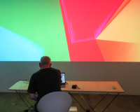 An image of a man, with his back turned to the camera, sat at a desk, manipulating an unseen controller for multi coloured lights and shapes projected onto the wall in front of him. The shapes are a warm mix of green, red, orange and yellow.