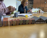 Two people sit at a table assembling small, colorful paper model houses. The atmosphere is focused and creative, with a variety of materials spread out.