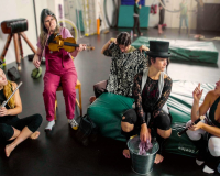 A group of circus performers, all white women, sit in a semi circle in a rehearsal space with a black floor. Some sit on the floor, while others sit on green cushions or chairs. They are in discussion, while two performers on the left play instruments - one, on the far left, plays the flute, while the other to her right plays a fiddle. 