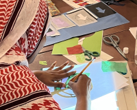 A person in a red and white patterned headscarf creates art with paper and scissors on a lightbox at a craft table, conveying focus and creativity.