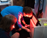 Two people examine a small cabinet under a bright red light in a dim room. One person opens a drawer, while the other watches, both appearing focused.