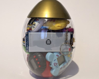 A transparent egg-shaped container with a gold top holding small toys, including a digital pet keychain and a smiling emoji screen, against a plain background.