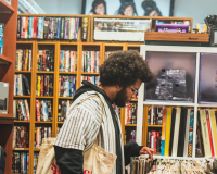 An image of the musician Lawi Anywar a Black man with an afro is browsing vinyl records in a vintage market. He is surrounded by various types of physical media, and carries a white tote bag full of records. 