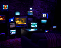An image of a darkened room, illuminated only by a series of CRT TV's organised in a disparate fashion, all activated and showing different images, creating a collage of sorts. There is a sofa placed in front of the installation, for the viewer to experience it. 