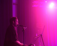 An image of Nik Rawlings singing into a microphone. There is a laptop in front of them, and the room they are performing in is illuminated entirely in a pinkish, purple hue. Nik wears all black and is prominent to the left of the image. 