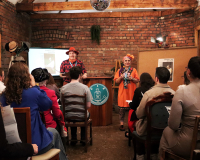 An image depicting Paul (on the left in red) and Gill (on the right in orange) delivering a theatre experience in a red brick room with a smartly dressed audience. They are flanked by a cork board on the right and a small projection set upon the left. 