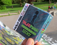 Thumb and fingers hold a computer game case saying 'Solar Punk Bristol Lucky' in the foreground, with a park and skate park in the background