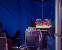 An image of men watching TV in a refugee camp. The TV shows a football match. 