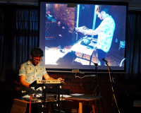 Ant Nasce a white man with glasses on stands at a Korg synthesizer, performing music under dim lighting. A large screen behind him displays a projected image of another person adjusting equipment, conveying a creative and focused atmosphere.