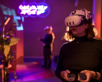 Person standing with a VR headset against a brightly coloured environment with another person in a headset in the background