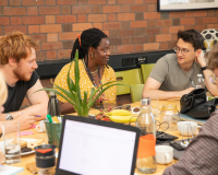 Five people engaged in a friendly discussion around a table with laptops and drinks. A plant adds greenery. The setting is casual and collaborative.