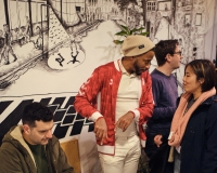 A man plays video games at a round table, while a person in a red jacket and beanie talks to a smiling woman. The background features a detailed mural.