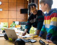 Smiling woman and boy collaborate at a table with a laptop and craft supplies in a bright classroom, creating a warm, engaging scene.