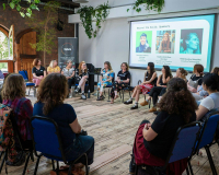 Group of people sitting in a circle looking at a person presenting with a screen behind them.