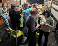 Adults gather in a cozy brick-walled workshop with string lights, chatting around tables and chairs; a screen and colorful artwork create a creative mood.